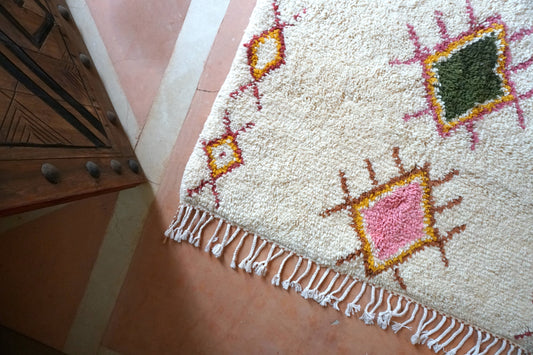 Close-up, overhead view of a white Moroccan wool rug with pink and green diamonds on top of a pink concrete floor with white inlay next to a carved wooden door with metal accents