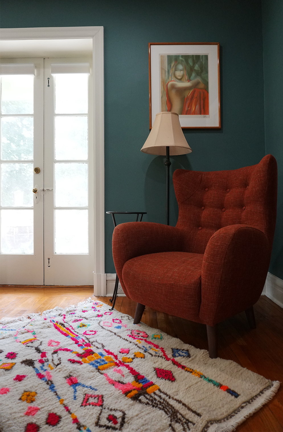 Red armchair in a room with a colorful rug, lamp, and framed picture on the wall.